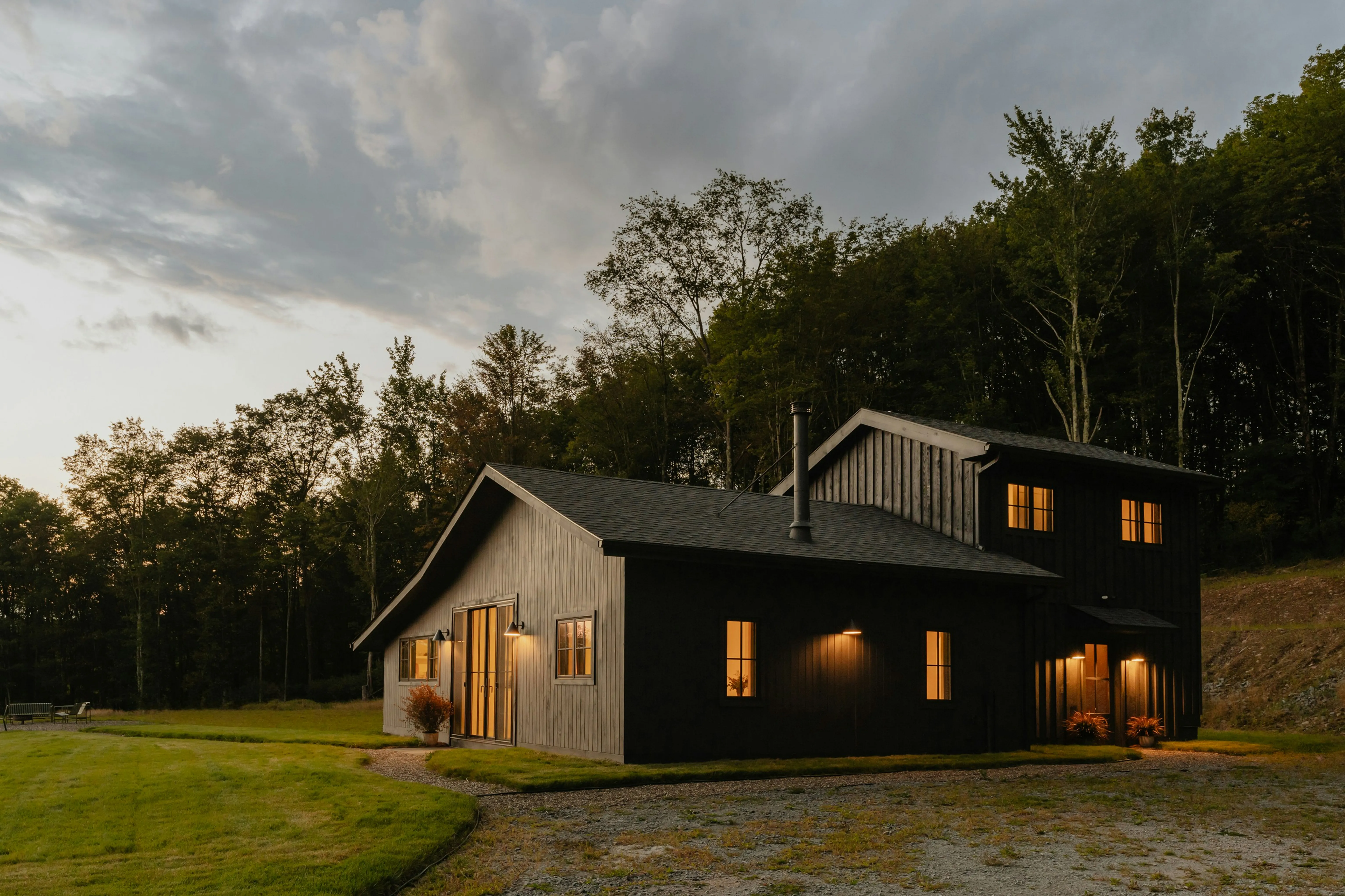 A modern barn-style home at dusk, its windows lit from within, set against a treeline above a gravel drive.